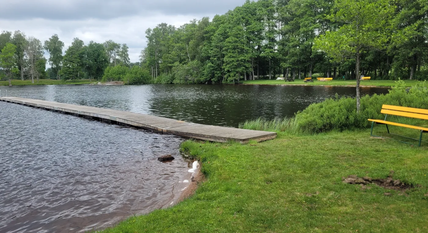 En brygga ut i Bjädesjöbadet med en gul bänk på stranden.