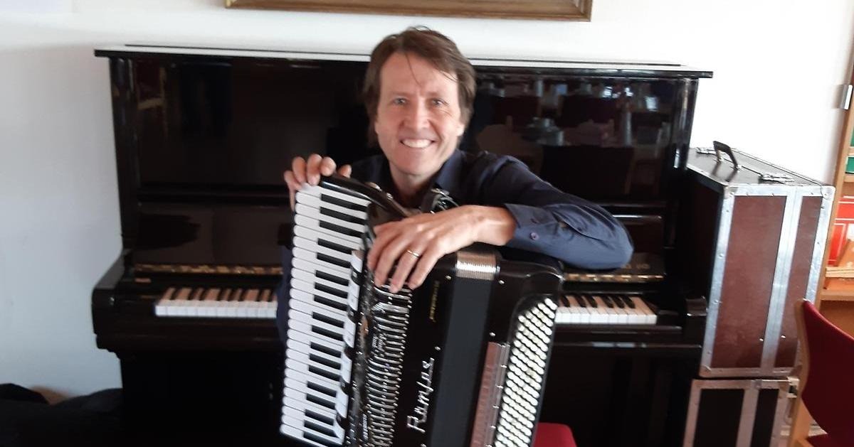 En man sitter med ett dragspel i knät, framför ett piano.
