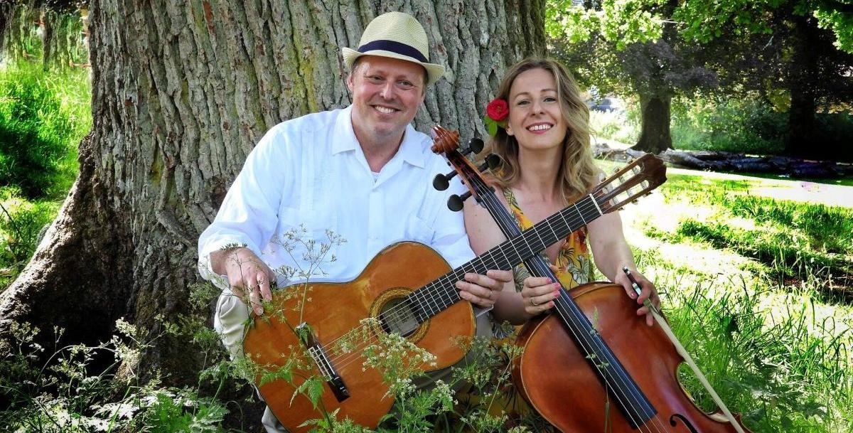 En man och en kvinna sitter i gräset framför ett träd, med gitarr och cello.