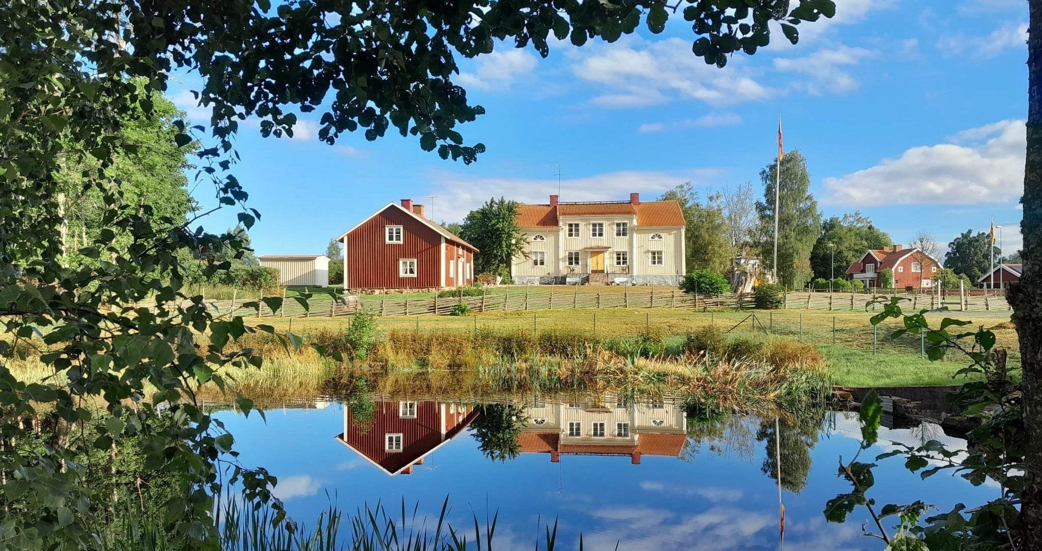 Bilden visar två byggnader som tillhör Björkö prästgård som speglar sig i en damm omgivet av lummig grönska