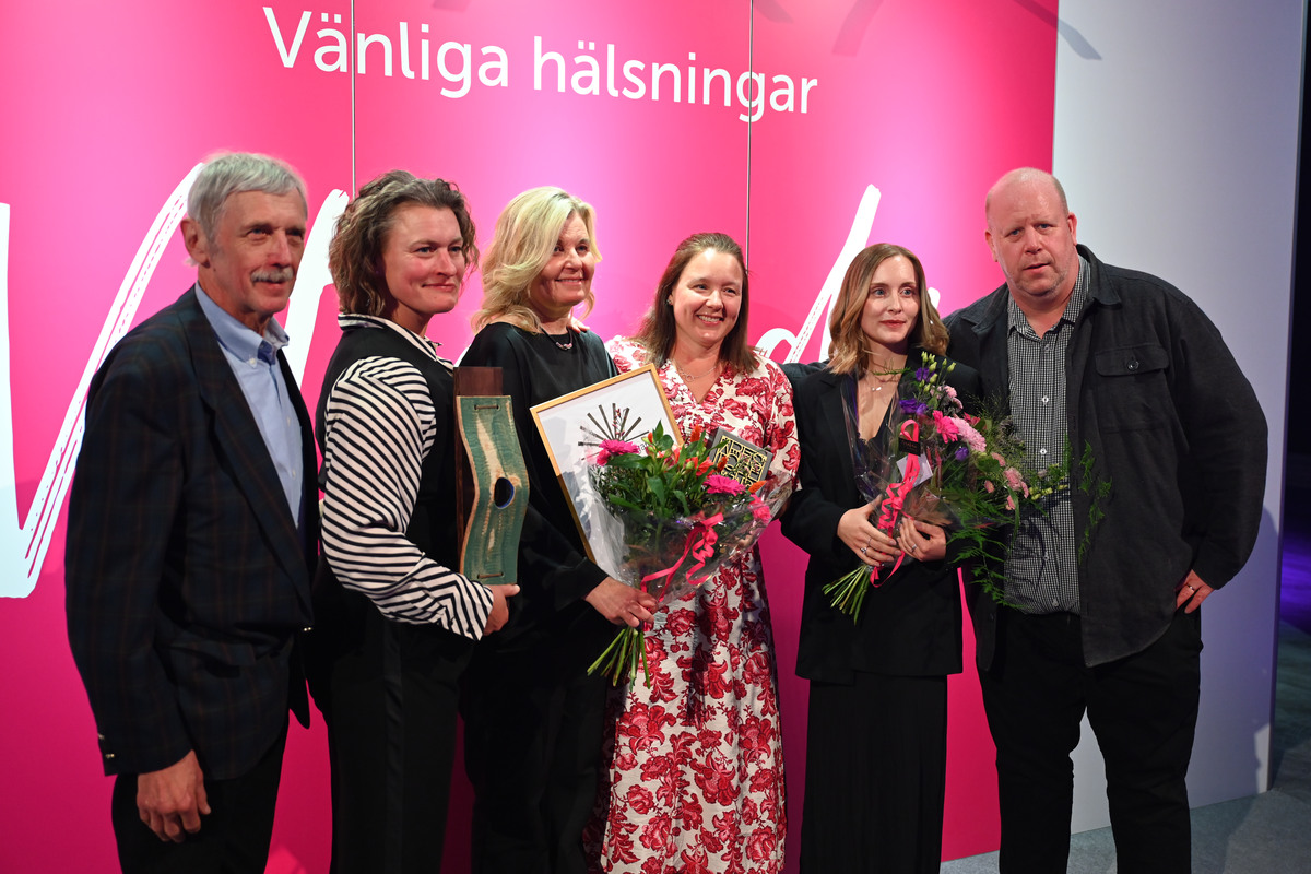 Två män och fyra kvinnor håller i statyett, diplom och blombukett framför en fotovägg.