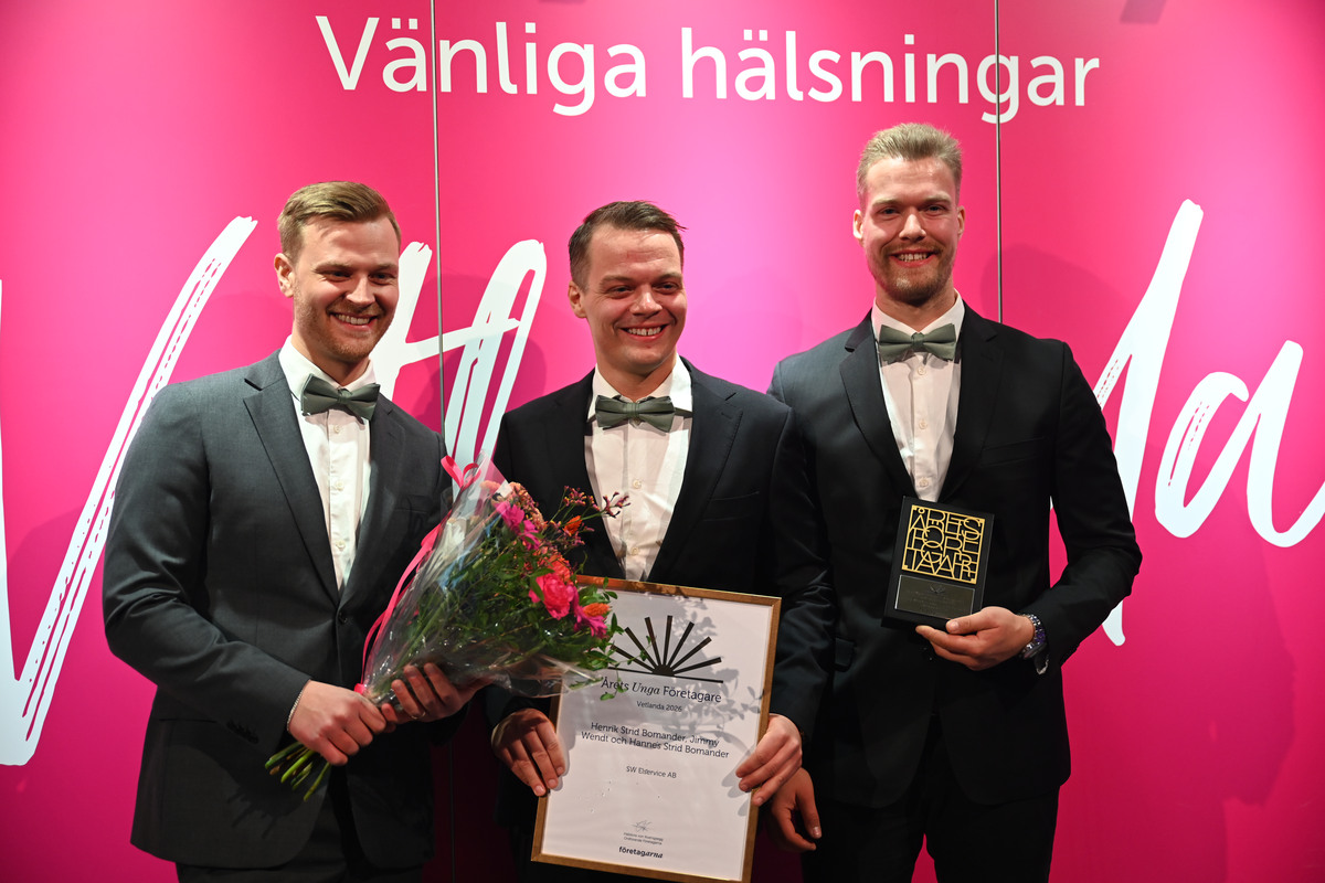 De tre killarna bakom SW Elservice står framför en fotovägg med blombukett, diplom och en statyett.
