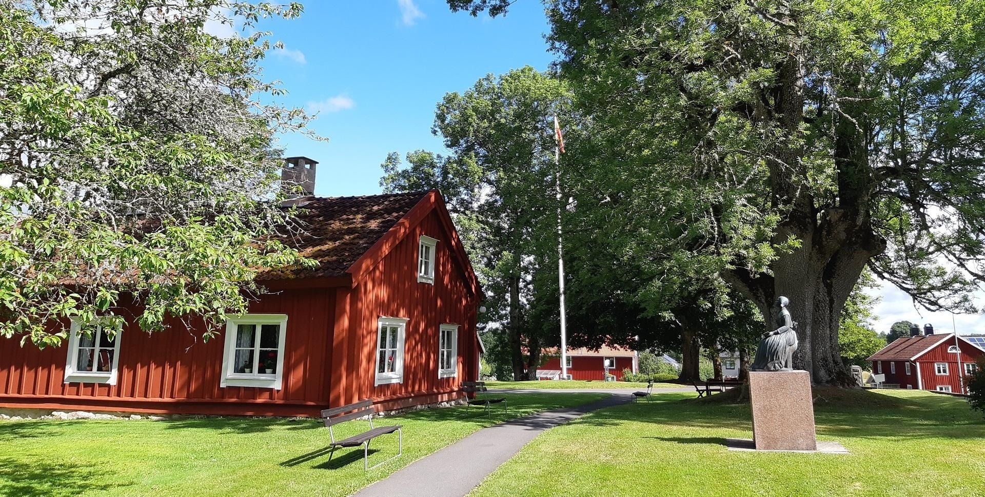 En röd stuga bland grönskande träd, en stor ask och statyn av Lina Sandell. Solen skiner.